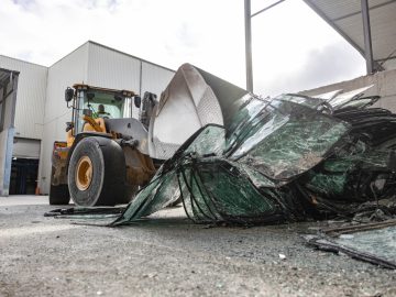 We zien een front-end-lader gebroken glasstukken opscheppen in een industriële omgeving met magazijnen op de achtergrond, terwijl een Audi Q4 e-tron voorbij komt, die een mix van ruige industrie en gestroomlijnde auto-innovatie laat zien.