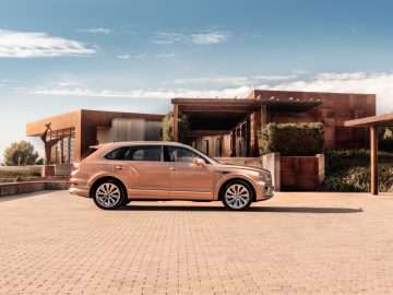 Een bronzen Bentley Bentayga SUV, in wezen een privéjet op wielen, staat geparkeerd voor een modern, minimalistisch huis met een plat dak en grote ramen. Het huis is omgeven door groen en de lucht is helder.