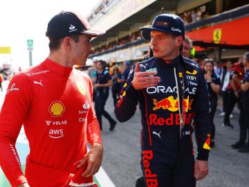 Twee Formule 1-coureurs in teamuniformen, de een in het rood en de ander in het blauw, praten terwijl ze door de pitstraat van een racecircuit lopen. Met teamleden en ander personeel op de achtergrond bespreken ze strategieën en lachen ze mogelijk zelfs met elkaar om de recente race van Max Verstappen, terwijl Drake's nieuwste hit vlakbij speelt.