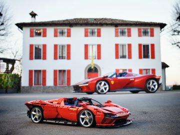 Een LEGO Ferrari-model staat op de weg voor een echte rode Ferrari, met een wit gebouw met rode luiken op de achtergrond.