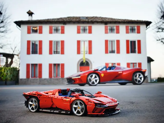 Een LEGO Ferrari-model staat op de weg voor een echte rode Ferrari, met een wit gebouw met rode luiken op de achtergrond.