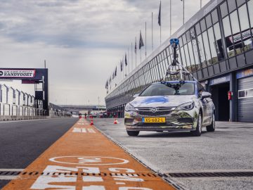 Een Street View-auto uitgerust met een hoge camera-opstelling rijdt langs de pitstraat van een racecircuit, met moderne gebouwen en vlaggen op de achtergrond.