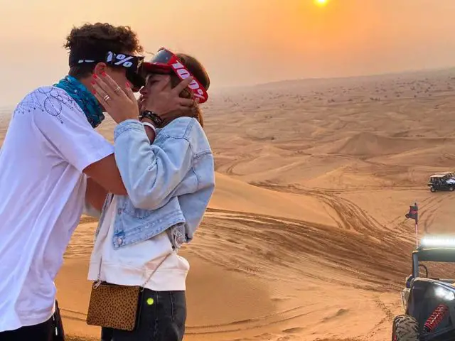 Een stel staat bij zonsondergang dicht bij elkaar in een woestijnlandschap, met bandana's en veiligheidsbrillen op, met terreinvoertuigen zichtbaar op de achtergrond, die de avontuurlijke geest van Lando Norris kanaliseren.