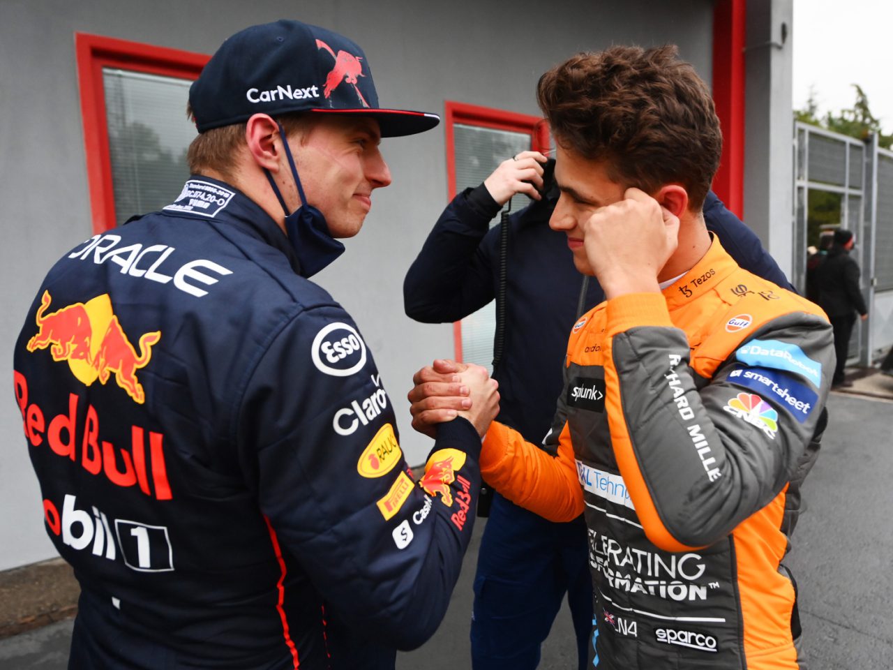 Twee Formule 1-coureurs in racepakken, Lando Norris in een McLaren-pak en een andere coureur in een Red Bull-pak, schudden elkaar de hand en glimlachen buiten een gebouw.