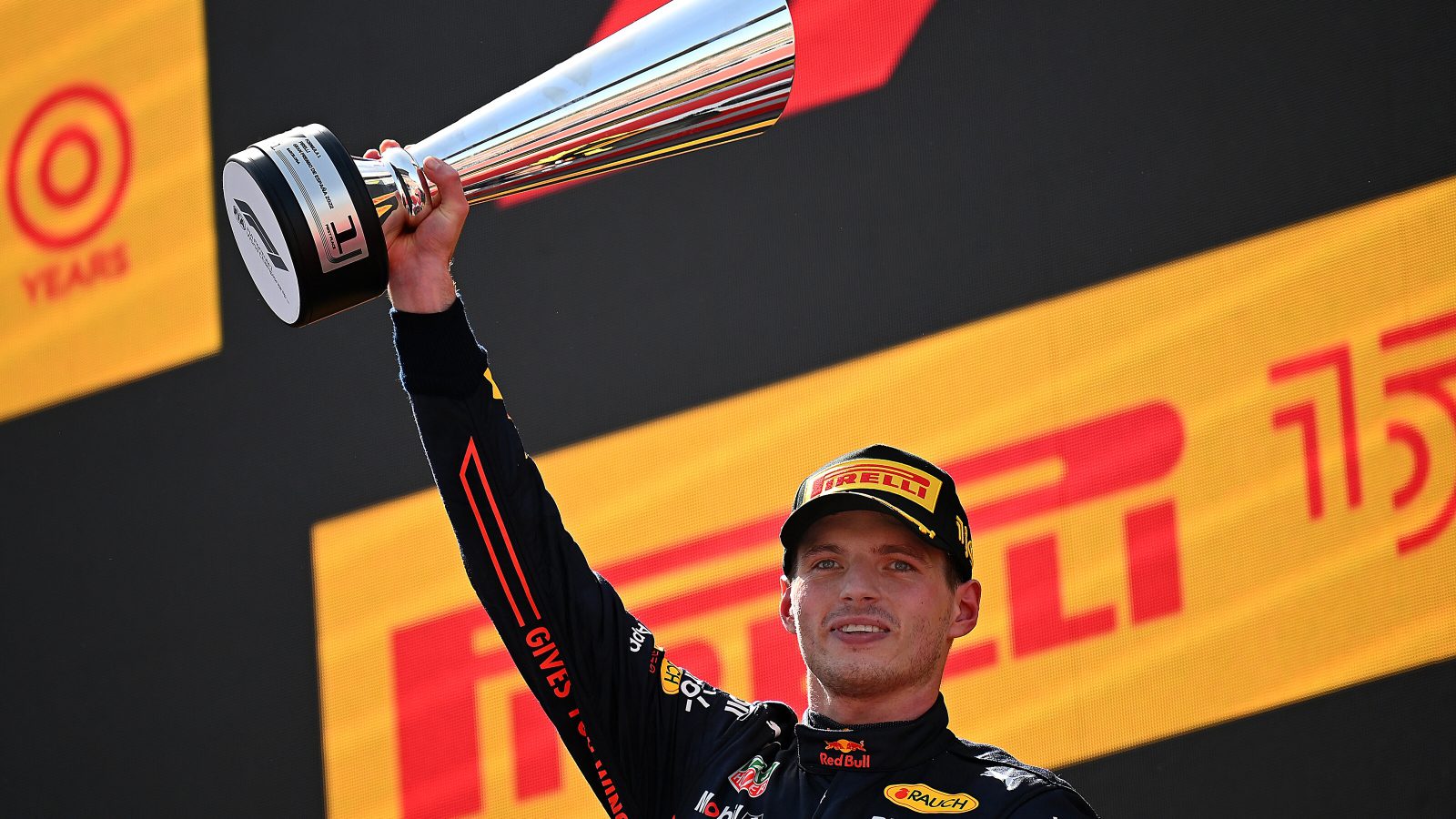 Een coureur houdt een grote zilveren trofee omhoog op een podium, die doet denken aan de overwinningsmomenten van Max Verstappen, met een pet op en een racepak versierd met logo's van sponsors.