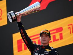 Een coureur houdt een grote zilveren trofee omhoog op een podium, die doet denken aan de overwinningsmomenten van Max Verstappen, met een pet op en een racepak versierd met logo's van sponsors.