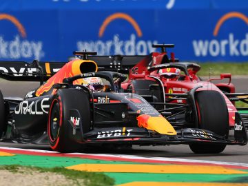 Twee Formule 1-auto's, de ene zwart met rode en gele accenten en de andere rood, racen nauwlettend op een circuit met 'Workday'-borden op de achtergrond tijdens de Grand Prix van Miami.