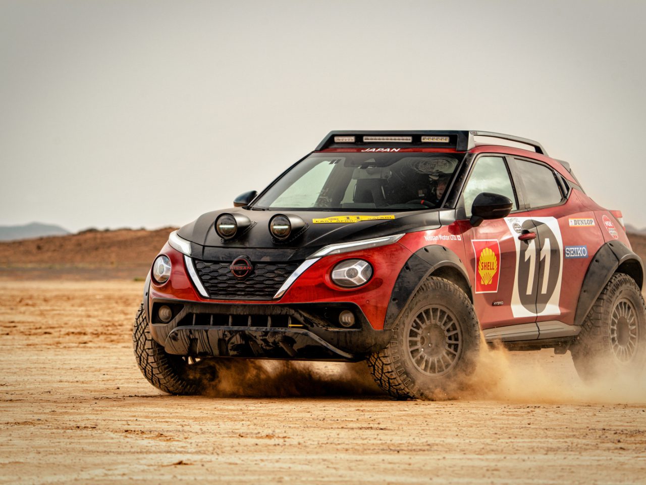 Een rode Nissan Juke Hybrid rallyauto met het nummer 11 op de zijkant rijdt tijdens een race door een stoffig woestijnterrein.