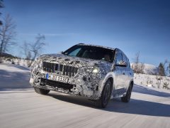 Een gecamoufleerde BMW X1 rijdt op een besneeuwde weg met een achtergrond van bomen en bergen onder een helderblauwe lucht.