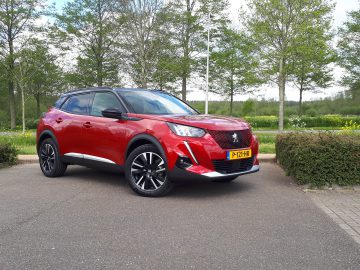 Een rode Peugeot e-2008 SUV geparkeerd op een verhard terrein met bomen en groen op de achtergrond, onder een gedeeltelijk bewolkte hemel.