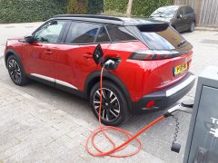 Een Peugeot e-2008, schitterend rood, is aangesloten op een laadstation in een woonwijk met een groene heg en houten hek op de achtergrond. Kentekenplaat gedeeltelijk zichtbaar.