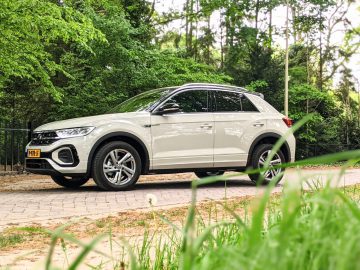 Een witte Volkswagen T-Roc SUV staat geparkeerd op een stenen pad in een bosrijke omgeving met weelderige groene bomen en gras op de achtergrond, wat het decor vormt voor een perfecte recensie.