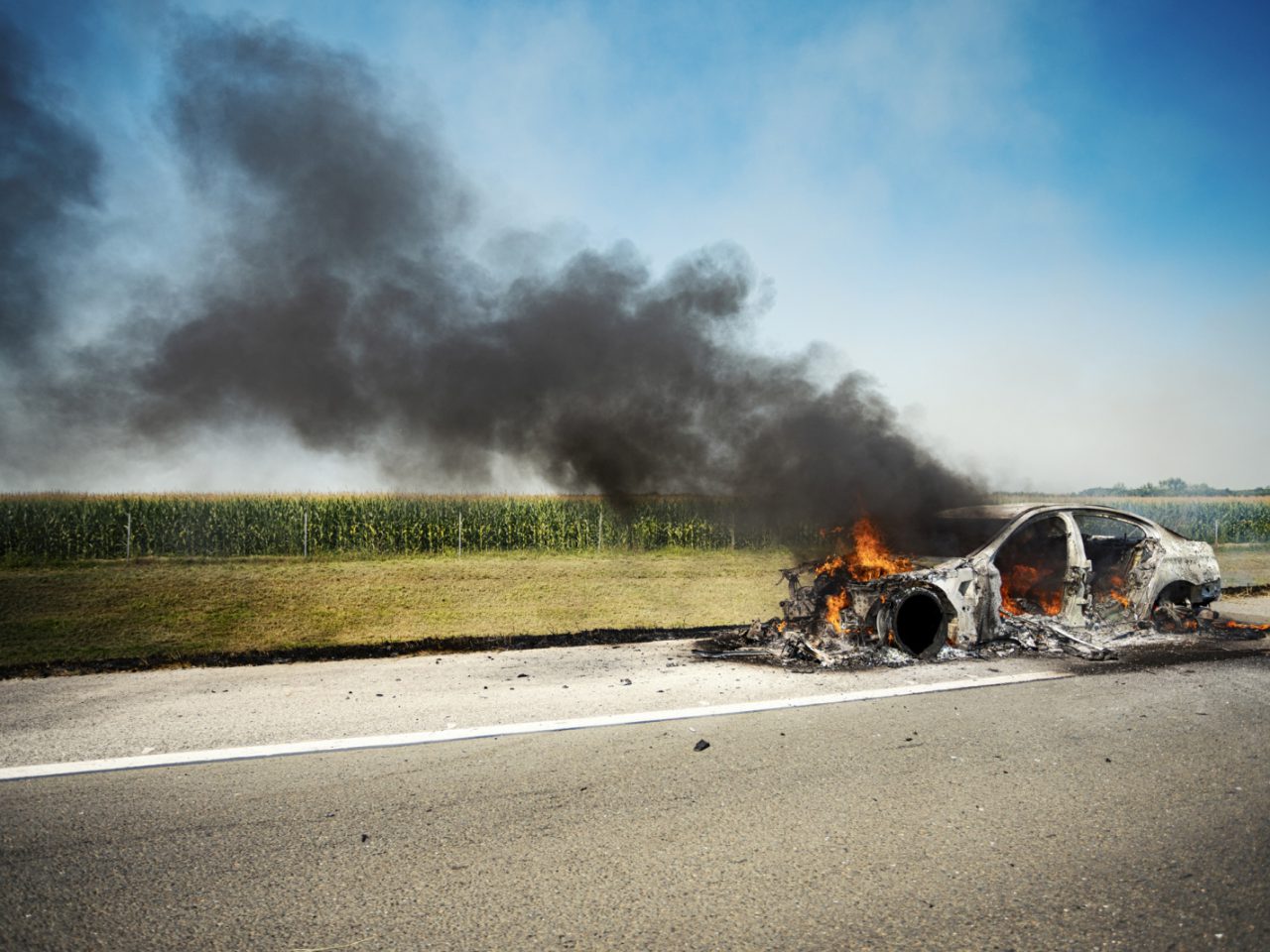 Een uitgebrande elektrische auto stoot dikke zwarte rook uit aan de kant van een landelijke weg, met een veld op de achtergrond.