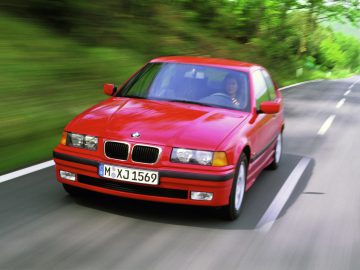 Een rode BMW E36-auto rijdt op een tweebaansweg omgeven door groen, met een onscherpe achtergrond die beweging aangeeft.