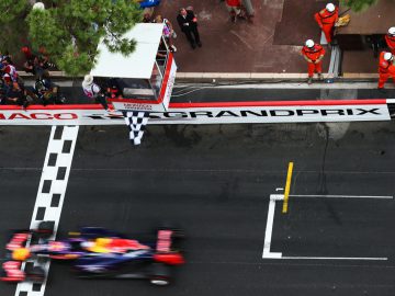 Een Formule 1-auto snelt langs de finish tijdens de Grand Prix van Monaco, terwijl officials in oranje uniformen en toeschouwers gretig toekijken, terwijl Formule 1-vlaggen op de achtergrond wapperen.