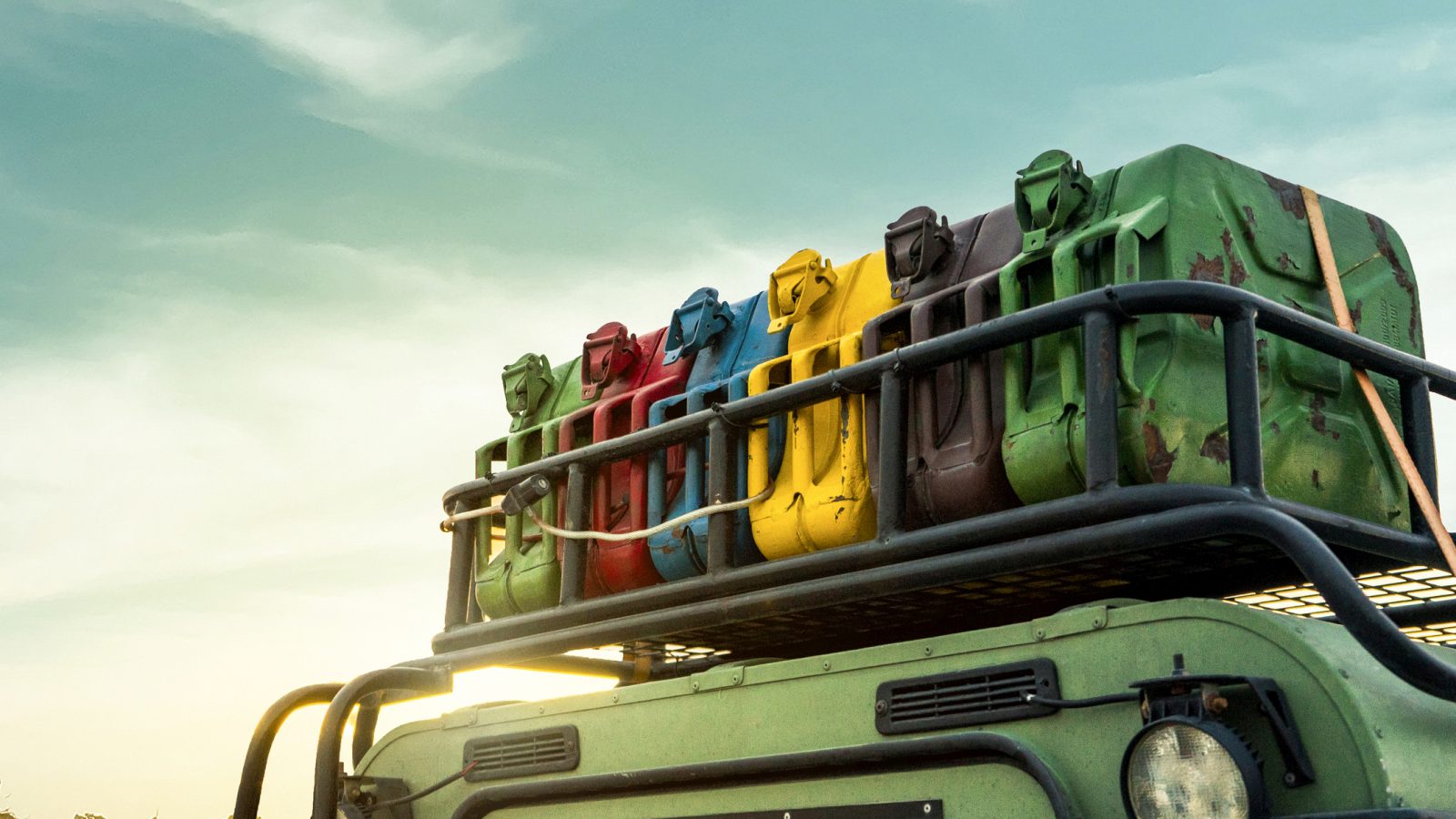 Dak van een groen voertuig met zes kleurrijke jerrycans gemonteerd op een imperiaal tegen een zonsondergang op de achtergrond, wat de sfeer van benzinereizen door het pittoreske Deutschland oproept.