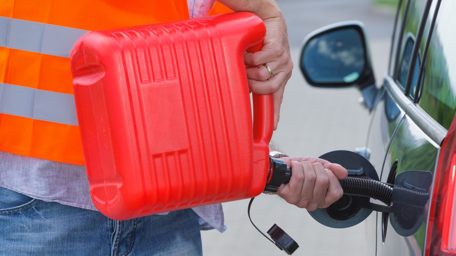 Een persoon in een reflecterend hesje tankt een auto met een rode gasfles gevuld met benzine, wat doet denken aan de ordelijke tankstops in Duitsland.