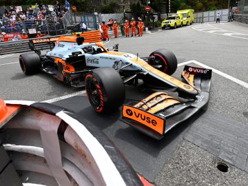 Een Formule 1-auto, omringd door veiligheidsbarrières, hulpverleners en een ambulance, staat geparkeerd op het iconische racecircuit van Monaco tijdens een openbaar evenement met toeschouwers op de achtergrond.