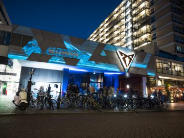 Nachtelijk stadsbeeld: een gebouw met een modern geometrisch ontwerp en verlichte bewegwijzering, met een stijlvolle Cupra-garage. Fietsen en scooters geparkeerd voor de deur. Hoogbouw zichtbaar op de achtergrond. Buiten verzamelden zich mensen.