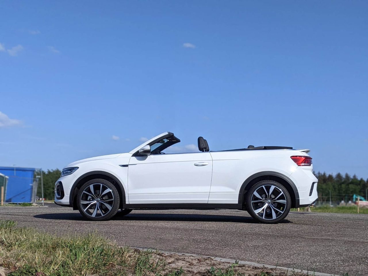 Een witte Volkswagen T-Roc Cabrio staat met de kap naar beneden geparkeerd op een verharde weg. De achtergrond toont een blauwe lucht, een grasveld en bomen.