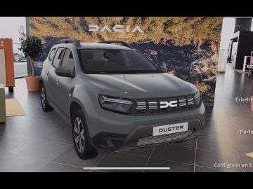 In een showroom staat een grijze Dacia Duster tentoongesteld. Op de achtergrond staat het prominente Dacia-logo en een ruig landschapsbeeld, wat zijn avontuurlijke karakter benadrukt. Ondertussen is aan de rechterkant handig een overlay van de gebruikersinterface zichtbaar.