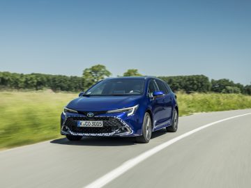 Een blauwe Toyota Corolla Hybrid rijdt op een gebogen weg met groen gras en bomen op de achtergrond onder een heldere hemel.