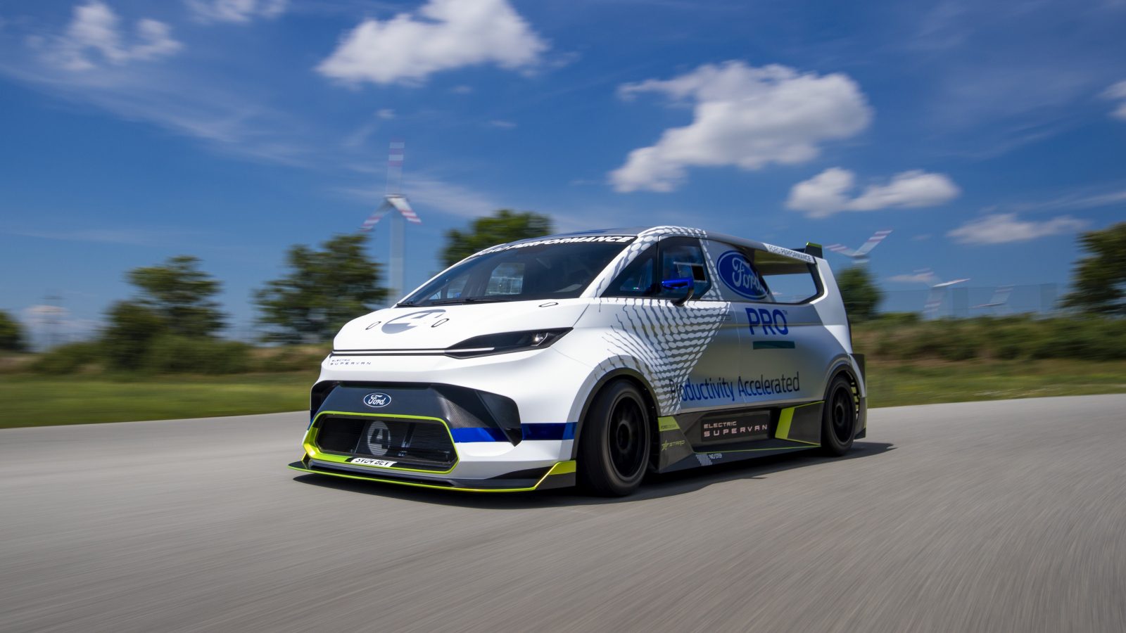 Een witte, aangepaste Ford Transit elektrische bestelwagen rijdt op een weg met een helderblauwe lucht en windturbines op de achtergrond.