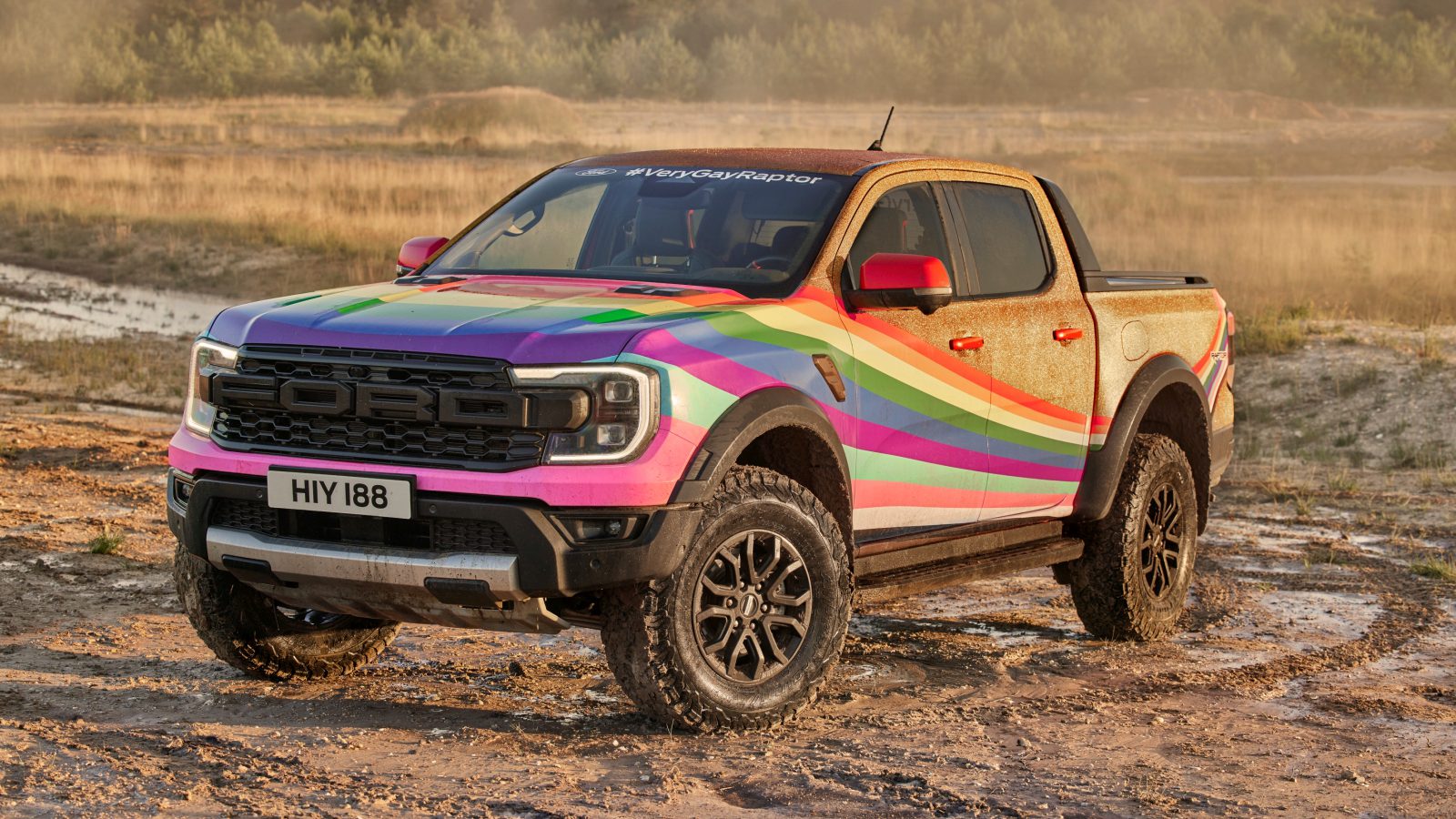 Een kleurrijke Ford Raptor pick-up met regenboogstrepen en de hashtag #VeryGayRaptor op de motorkap staat onder de modder geparkeerd op een ruig buitenterrein.