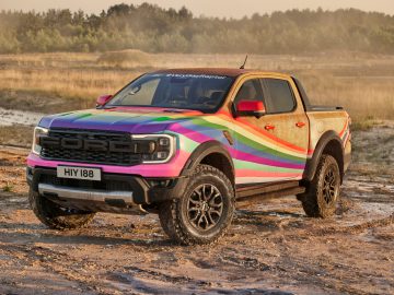 Een kleurrijke Ford Raptor pick-up met regenboogstrepen en de hashtag #VeryGayRaptor op de motorkap staat onder de modder geparkeerd op een ruig buitenterrein.