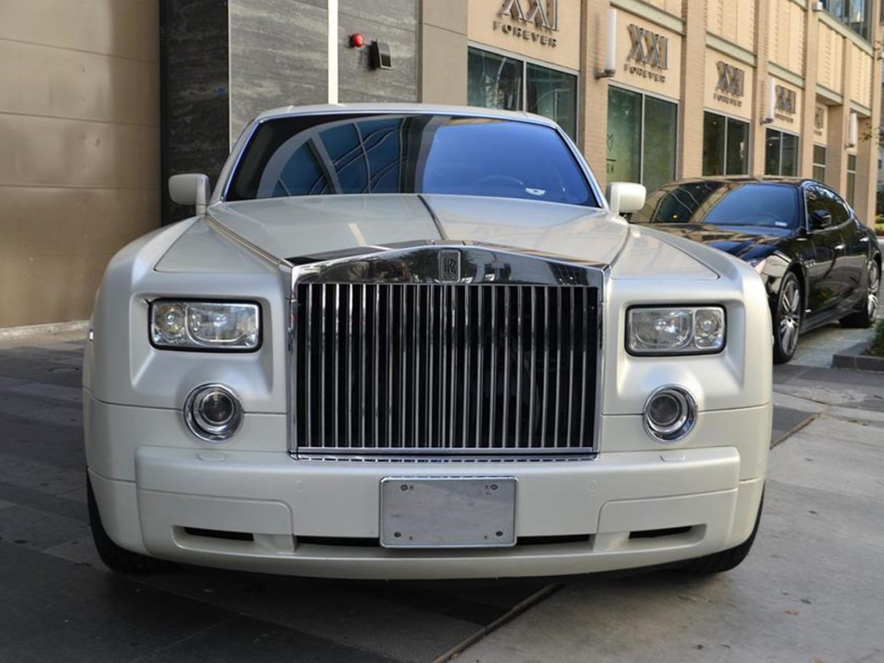 Een witte luxe auto met een prominente grille en rechthoekige koplampen staat geparkeerd in een stadsstraat en biedt de smaak van goedkoop autorijden, met een zwarte auto zichtbaar op de achtergrond.