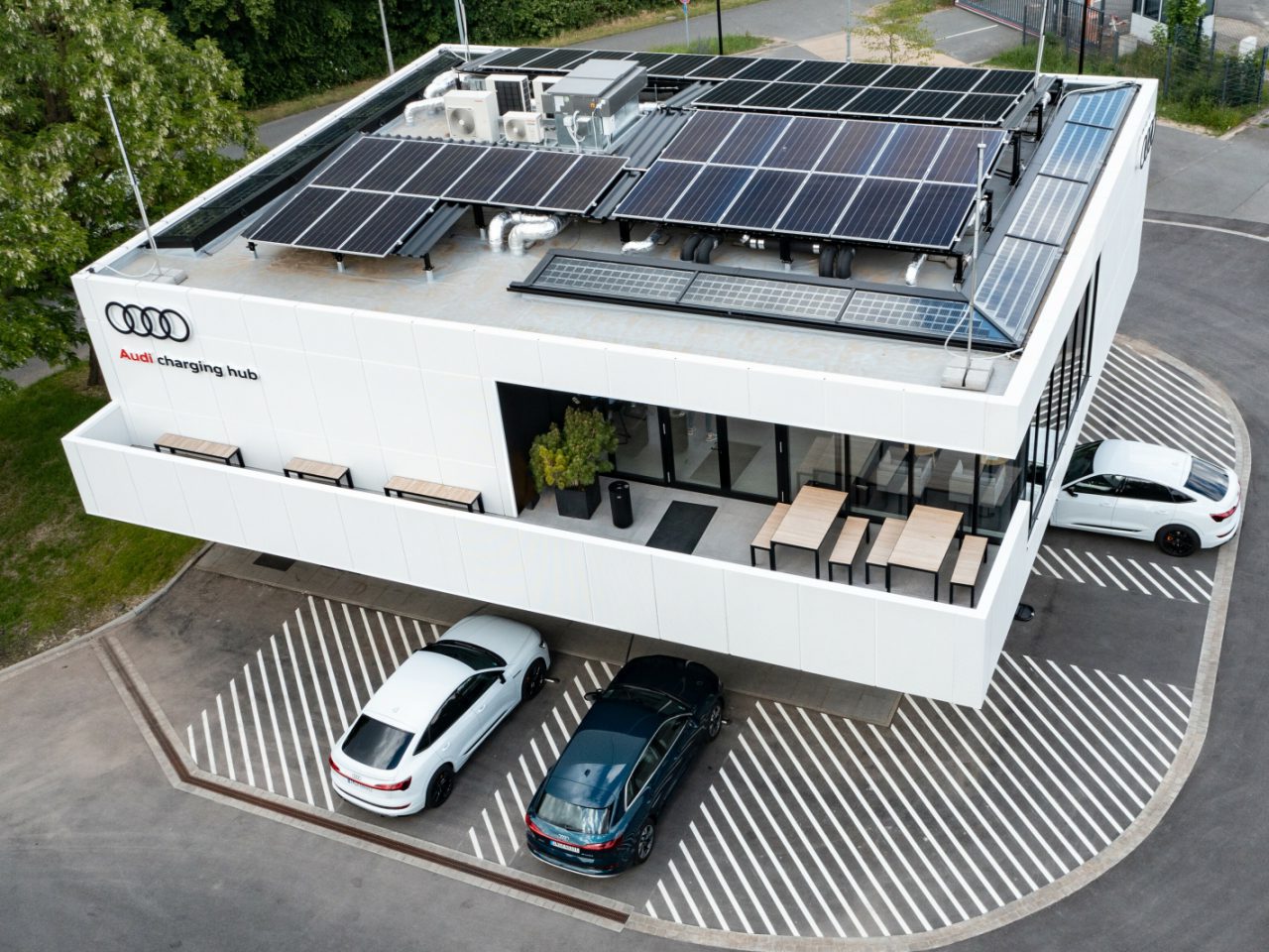 Luchtfoto van een modern gebouw met zonnepanelen op het dak, met het opschrift 'Audi-oplaadhub'. Drie auto's staan geparkeerd op speciale parkeerplaatsen voor auto's met gestreepte markeringen rondom het gebouw.