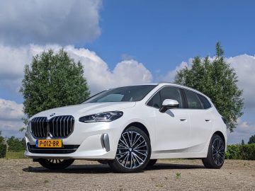 Op een verhard terrein staat een witte BMW 2 Serie Active Tourer met geel kenteken geparkeerd, met bomen en een bewolkte lucht op de achtergrond.