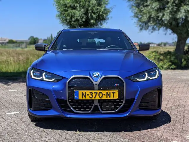 Een blauwe BMW-auto met koplampen aan, voorzien van een Nederlands kenteken N-370-NT, buiten geparkeerd op een zonnige dag. Perfect voor iedereen die het strakke ontwerp en de geavanceerde functies van de BMW i4-serie overweegt.