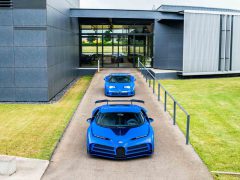 Twee blauwe sportwagens staan geparkeerd voor een modern gebouw met grote ramen en grijze buitenmuren. Zien: dit is de eerste klantgeleverde Bugatti Centodieci, die een vleugje exclusiviteit aan de scène toevoegt.