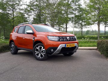 Een oranje Dacia Duster staat geparkeerd op een verhard terrein voor een graslandschap met bomen op de achtergrond. Het voertuig heeft een Europees kenteken.