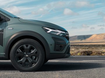De voorkant van een groene Dacia SUV, voorzien van de nieuwste facelift, is geparkeerd op een verharde weg met een landschap van heuvels en een bewolkte lucht op de achtergrond.