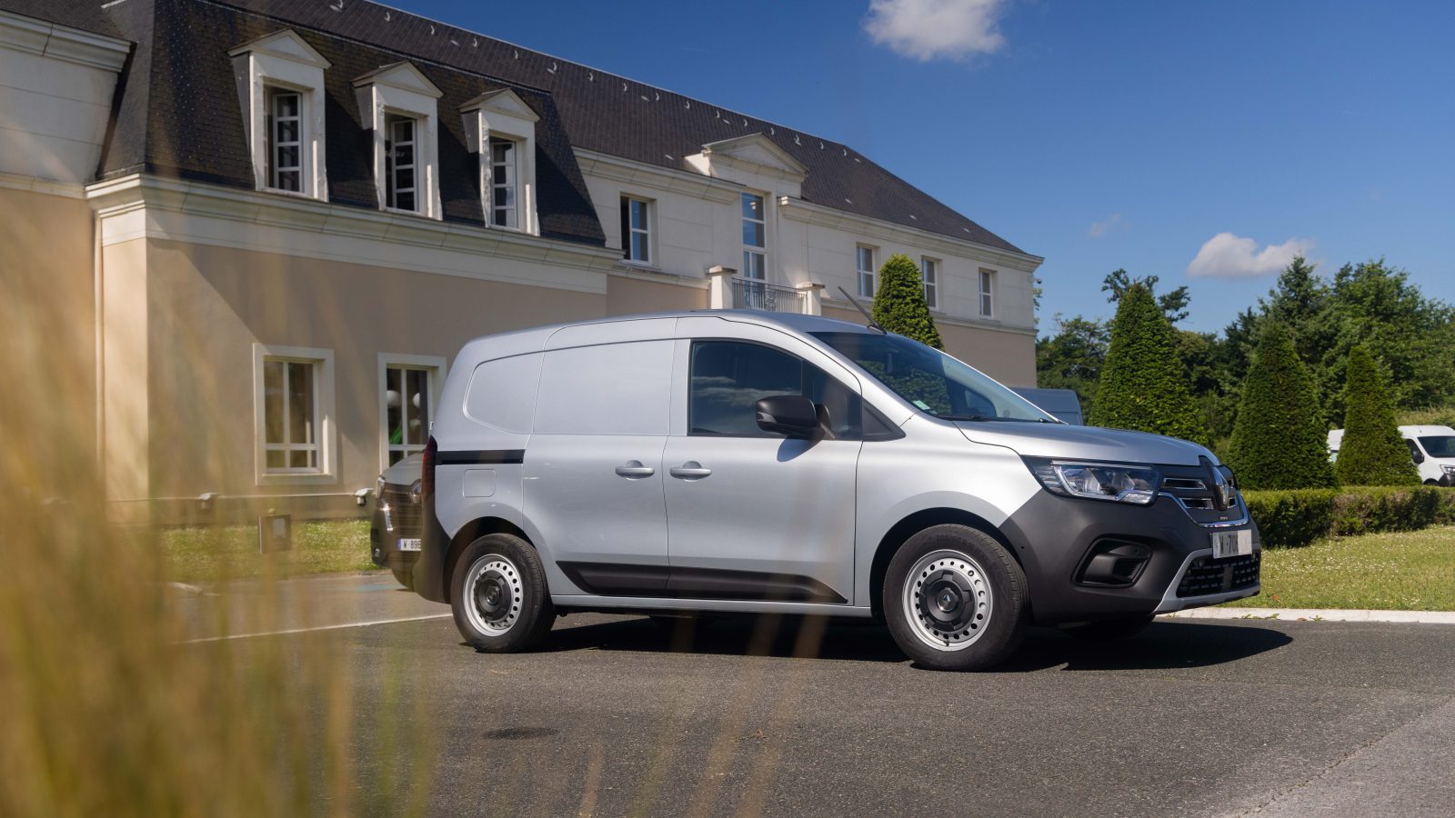 Een elektrische bestelwagen van Renault Master E-Tech staat geparkeerd voor een groot beige gebouw met donkere dakpannen op een zonnige dag, met bomen en een blauwe lucht op de achtergrond.