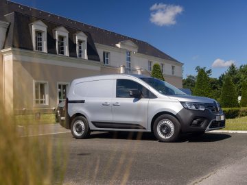 Een elektrische bestelwagen van Renault Master E-Tech staat geparkeerd voor een groot beige gebouw met donkere dakpannen op een zonnige dag, met bomen en een blauwe lucht op de achtergrond.