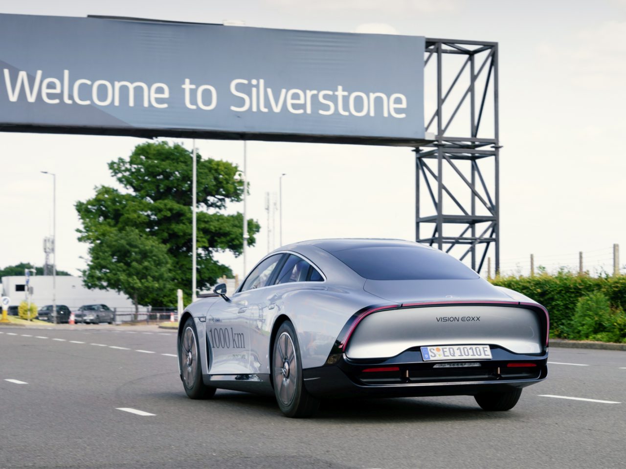 Een slanke, zilverkleurige auto rijdt op een weg met een groot bord erboven met de tekst 'Welkom in Silverstone'. De Mercedes elektrisch is op de achterzijde gemarkeerd met "VISION EQXX". Op de achtergrond zijn bomen en een hek zichtbaar.