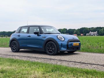 Een blauwe Mini Cooper met Nederlands kenteken staat geparkeerd op een landelijke weg met groene velden en bomen op de achtergrond en ziet eruit alsof hij zojuist uit een Mini review 2022-catalogus is gereden.
