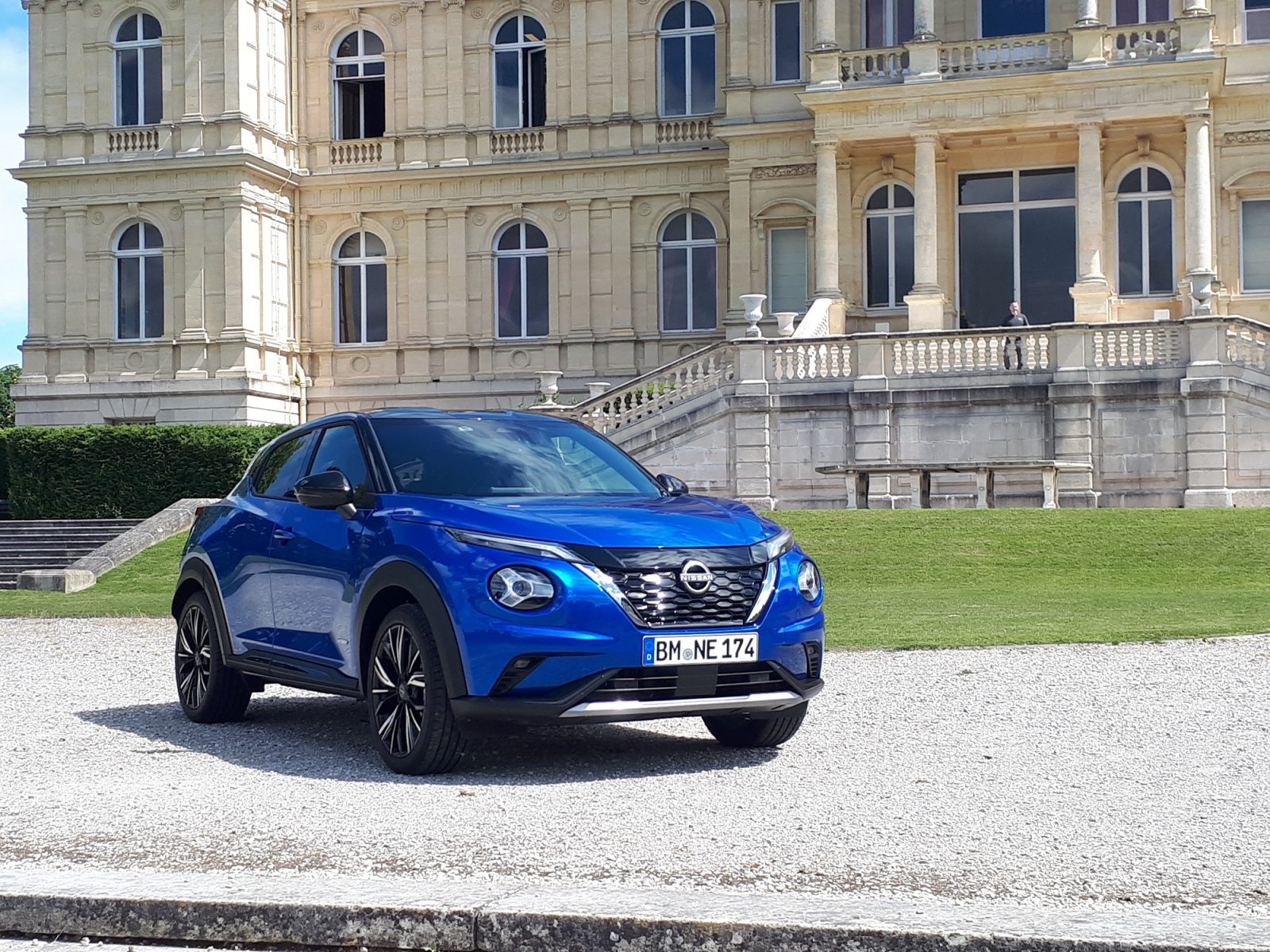 Een blauwe Nissan Juke Hybrid staat geparkeerd op een grindpad voor een groots, historisch gebouw met beige stenen muren en hoge ramen.