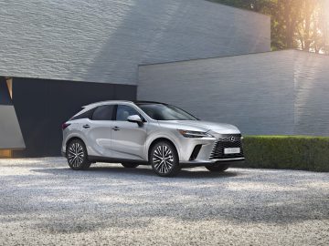 Een nieuwe Lexus RX, glanzend in zilver, staat geparkeerd op een grindoprit voor een modern gebouw met stijlvolle grijze bakstenen muren.