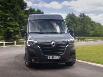Een grijze elektrische bestelwagen Renault Master E-Tech rijdt op een weg met groen en bomen zichtbaar op de achtergrond.