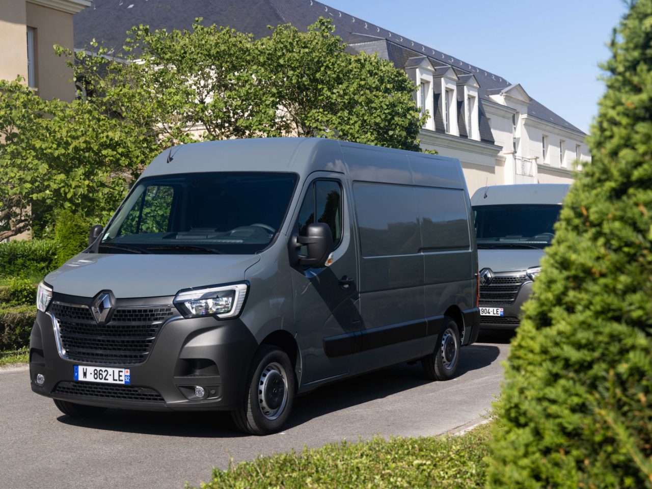 Twee grijze Renault Master E-Tech elektrische bedrijfswagens staan geparkeerd op een oprit omringd door groene struiken en bomen nabij een gebouw met een donker dak.