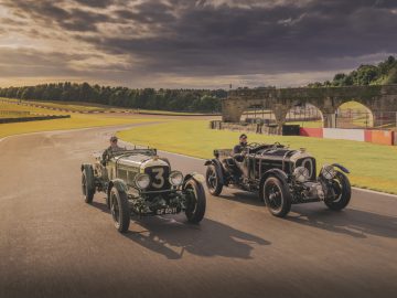 Twee vintage raceauto's, een groene en een zwarte Bentley, racen zij aan zij op een racecircuit met een gebogen stenen brug en groen op de achtergrond onder een bewolkte hemel.
