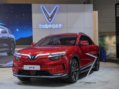 Een opvallende rode VinFast VF 8 SUV wordt tentoongesteld in een goed verlichte overdekte tentoonstellingsruimte, compleet met een groot scherm en prominente VinFast-branding op de achtergrond.