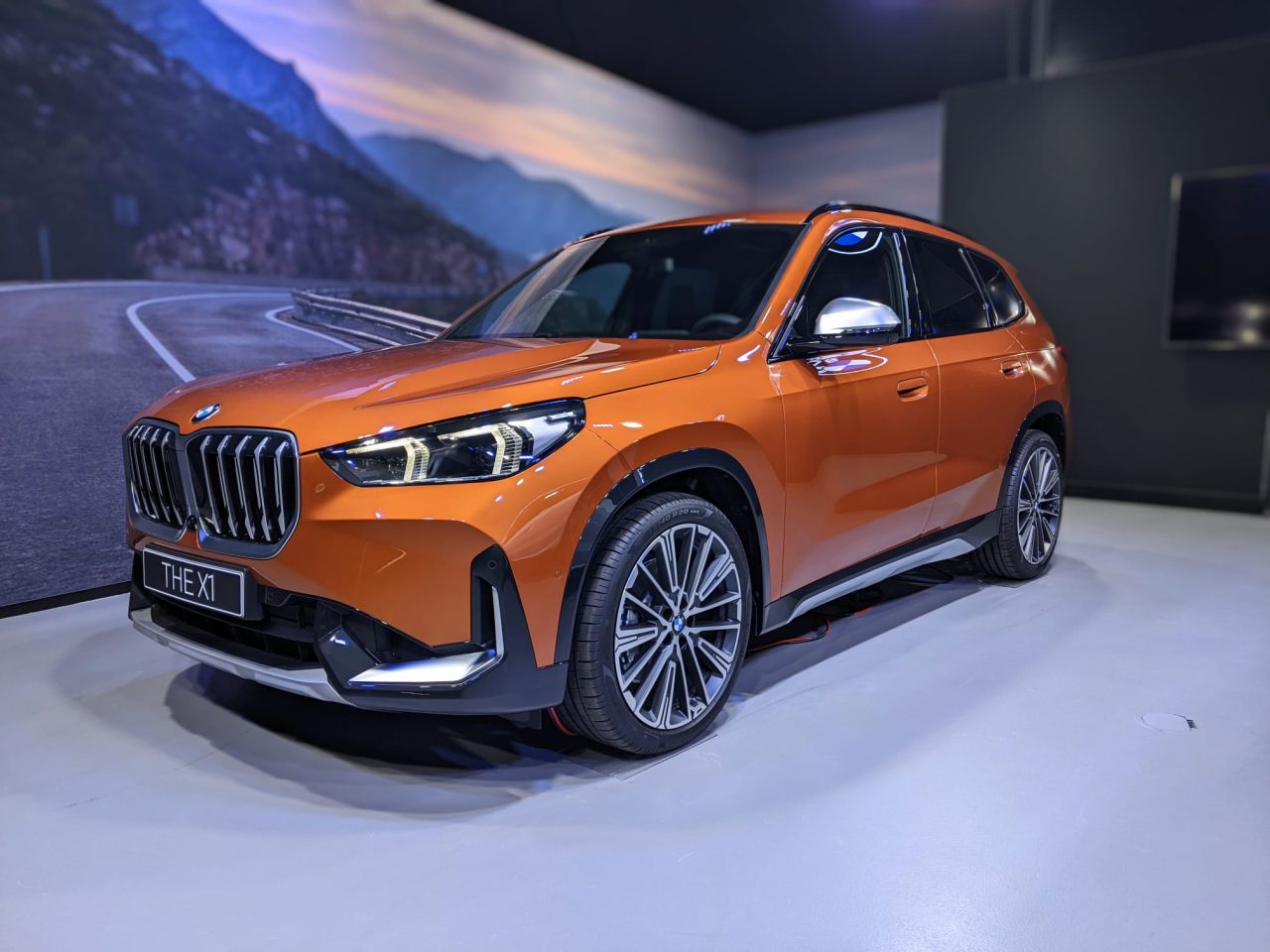 Een feloranje BMW X1 wordt binnenshuis tentoongesteld op een lichtgekleurde vloer, met een schilderachtige bergweg op de achtergrond die het tafereel versterkt.