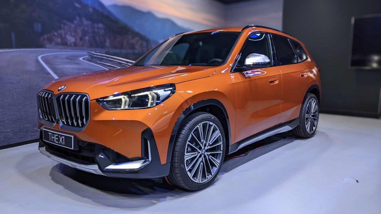 Een feloranje BMW X1 wordt binnenshuis tentoongesteld op een lichtgekleurde vloer, met een schilderachtige bergweg op de achtergrond die het tafereel versterkt.