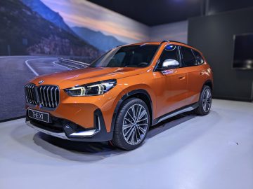 Een feloranje BMW X1 wordt binnenshuis tentoongesteld op een lichtgekleurde vloer, met een schilderachtige bergweg op de achtergrond die het tafereel versterkt.