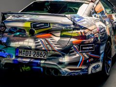 Binnen staat een auto geparkeerd met een kleurrijke wrap met daarop diverse afbeeldingen van racescènes, die doen denken aan de 3.0 CSL Hommage. Op het kenteken staat